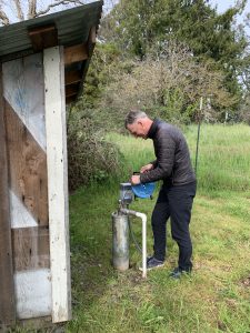 a man stands outside near a shed holding a blue device and measures his well