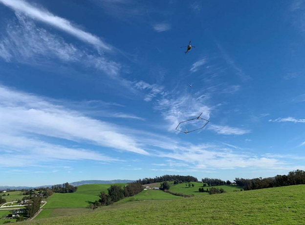Photo of blue sky and green rolling hills with a small helicopter in the air completing an AEM survey