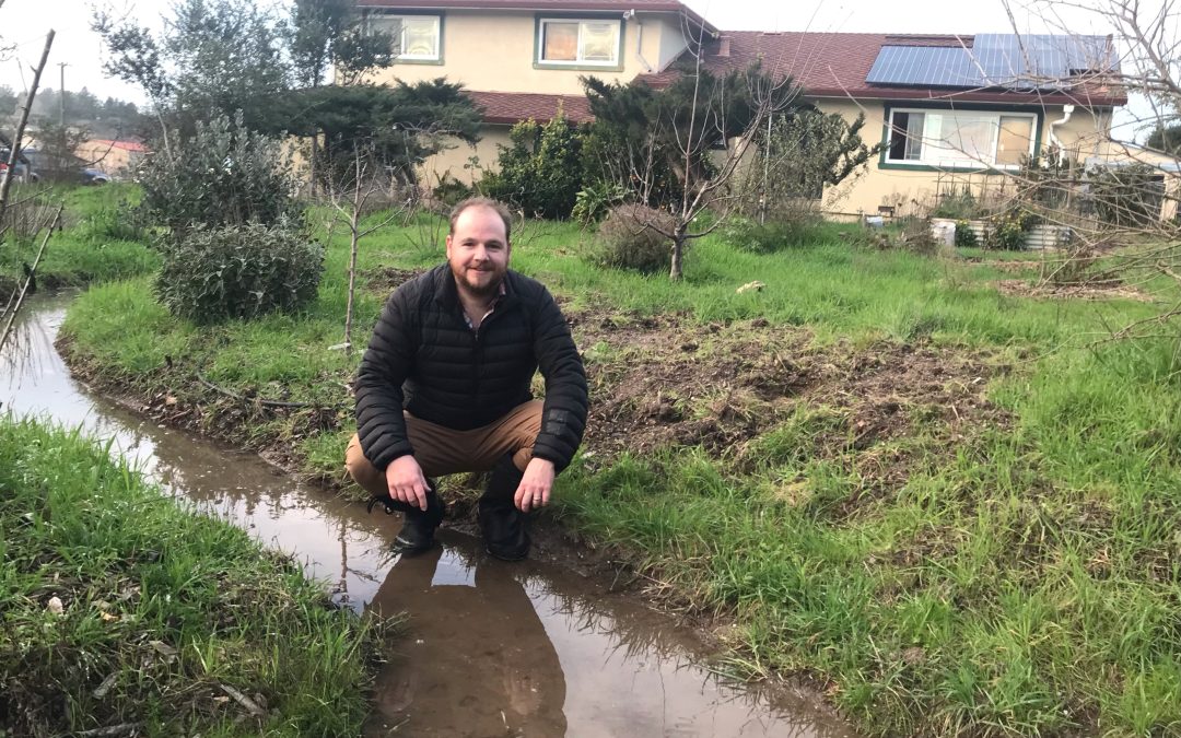 man crouches down in his yard next to a trench with water in it with his house in the background and green grass and plants around him